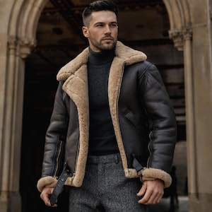 Pu&ograve; includere: Giacca in pelle marrone scuro con fodera e colletto in montone beige. La giacca presenta una chiusura a zip, un cinturino con fibbia e finiture in montone sui polsini. La persona indossa un dolcevita grigio e pantaloni grigi.