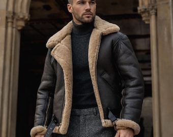 May include: A dark brown leather jacket with a beige shearling lining and collar. The jacket features a zip closure, a buckled strap, and shearling trim on the cuffs. The person is wearing a gray turtleneck and gray trousers.