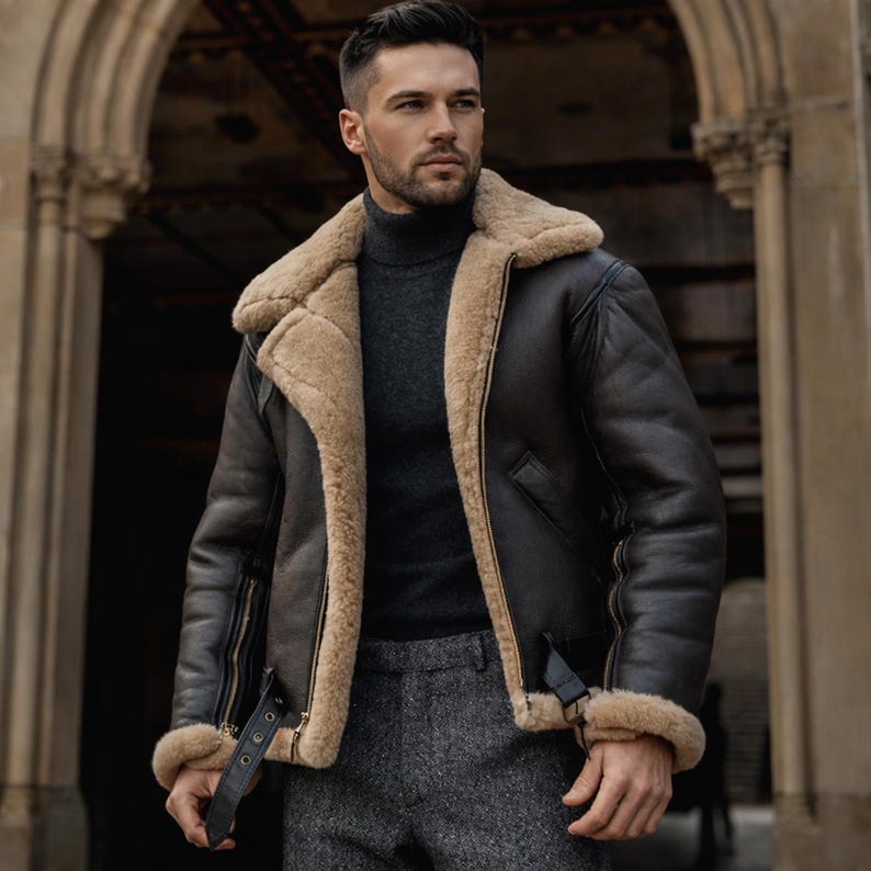 Pu&ograve; includere: Giacca in pelle marrone scuro con fodera e colletto in montone beige. La giacca presenta una chiusura a zip, un cinturino con fibbia e finiture in montone sui polsini. La persona indossa un dolcevita grigio e pantaloni grigi.