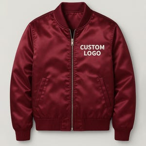 May include: A maroon satin bomber jacket with a full-length zipper closure and ribbed cuffs and hem. The jacket features the text "CUSTOM LOGO" in white block letters on the chest. The jacket has two front pockets and a classic bomber jacket design.
