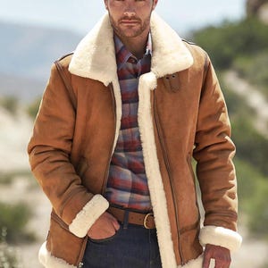 May include: A brown shearling jacket with a cream-colored shearling collar and trim. The jacket has a zipper closure, a brown leather belt, and is worn over a plaid shirt and blue jeans. The jacket's design is classic and warm.