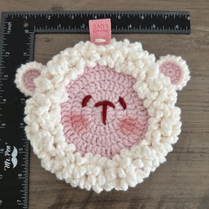 May include: A pink crocheted sheep with white yarn around the edges. The sheep has a small red nose and embroidered eyes. The sheep is about 4 inches in diameter. A pink tag with the text "Made with LOVE" is attached to the sheep.