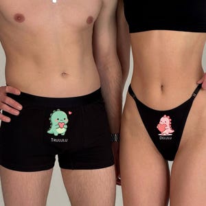 Op de afbeelding: Zwarte boxershorts en een zwarte string met bijpassende dinosaurusafbeeldingen. De boxershorts hebben een groene dinosaurus met een hart met het woord "TRULULU" eronder. De string heeft een roze dinosaurus met een hart met het woord "DELULU".