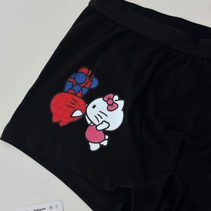 Pode incluir: Cuecas boxer pretas com um gr&aacute;fico colorido do Homem-Aranha e Hello Kitty. A roupa interior tem uma cintura el&aacute;stica preta e &eacute; feita de um material macio. Uma pequena etiqueta branca com texto tamb&eacute;m &eacute; vis&iacute;vel.