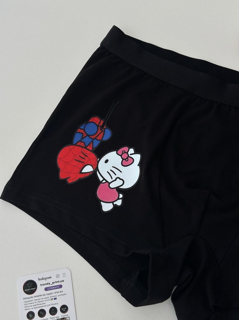 Pode incluir: Cuecas boxer pretas com um gr&aacute;fico colorido do Homem-Aranha e Hello Kitty. A roupa interior tem uma cintura el&aacute;stica preta e &eacute; feita de um material macio. Uma pequena etiqueta branca com texto tamb&eacute;m &eacute; vis&iacute;vel.