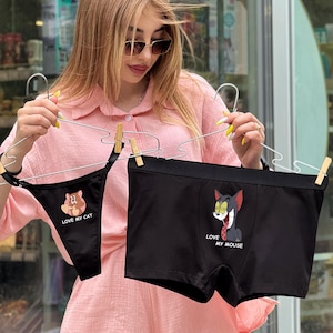 Op de afbeelding: Zwarte ondergoedset met cartoonafbeeldingen. Eén is een string met de tekst "Love My Cat" en een cartoonkat. De andere is een boxershort met de tekst "Love My Mouse" en een cartoonkat. Beide worden op hangers gehouden.