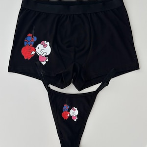 Pode incluir: Conjunto de roupa interior preta com boxers e fio dental. Ambas as pe&ccedil;as apresentam um gr&aacute;fico de desenho animado de um gato branco com la&ccedil;o rosa e cora&ccedil;&atilde;o vermelho, juntamente com um design vermelho e azul.