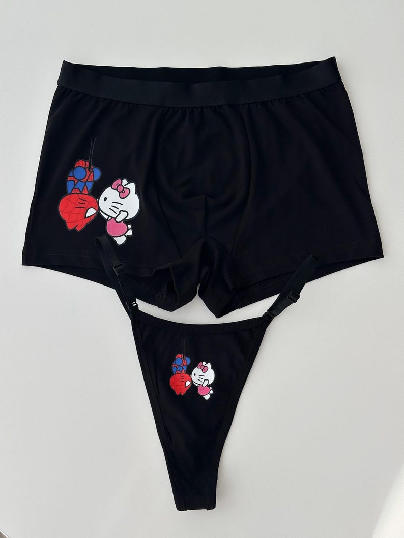 Pode incluir: Conjunto de roupa interior preta com boxers e fio dental. Ambas as pe&ccedil;as apresentam um gr&aacute;fico de desenho animado de um gato branco com la&ccedil;o rosa e cora&ccedil;&atilde;o vermelho, juntamente com um design vermelho e azul.