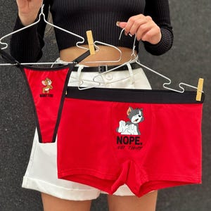 Könnte beinhalten: Rotes Unterwäsche-Set mit Zeichentrickfiguren. Eines ist ein String mit dem Text "WANT YOU" und Jerry Maus. Das andere ist eine Boxershorts mit Tom Katze und dem Text "NOPE. NOT TODAY". Beide werden an Kleiderbügeln gehalten.