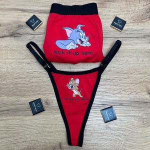 Könnte beinhalten: Rotes Unterwäsche-Set mit den Zeichentrickfiguren Tom und Jerry. Die Boxershorts zeigen Tom mit dem Text "suck it up here". Der String zeigt Jerry mit dem Text "for lick any whim". Schwarze Verzierungen und Träger.