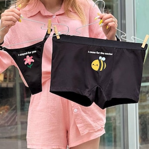 Könnte beinhalten: Schwarzes Unterwäsche-Set mit einem String-Tanga und Boxershorts. Der String-Tanga hat den Text "I stayed for you" und eine rosa Blumen-Grafik. Die Boxershorts haben den Text "I came for the nectar" und eine Cartoon-Biene.