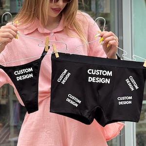 Può includere: Set di biancheria intima nera, composto da perizoma e boxer, entrambi con la scritta bianca "CUSTOM DESIGN". Gli indumenti sono tenuti da grucce. La persona nell'immagine indossa una camicia e pantaloncini rosa.