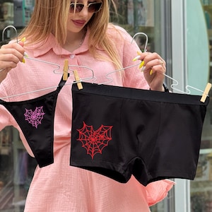 Op de afbeelding: Zwarte ondergoedset met een string en een boxershort, elk met een spinnenwebontwerp in roze en rood. De kledingstukken worden gepresenteerd op hangers met houten wasknijpers.