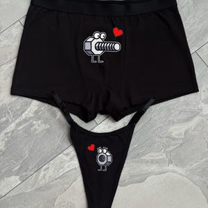 Puede incluir: Conjunto de bóxer negro y tanga a juego. Ambas piezas presentan un diseño de perno de dibujos animados con un corazón rojo. El conjunto de ropa interior está hecho de un material suave.