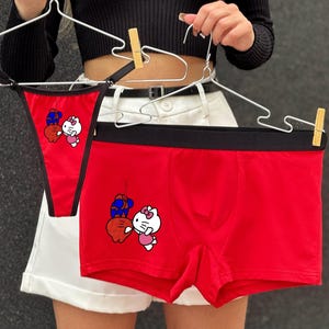Pode incluir: Conjunto de roupa interior vermelha com fio dental e cuecas boxer. Ambas as pe&ccedil;as t&ecirc;m uma cintura preta e um desenho animado de um gato branco com um la&ccedil;o rosa e um objeto azul e vermelho. A roupa interior est&aacute; pendurada em cabides de arame.