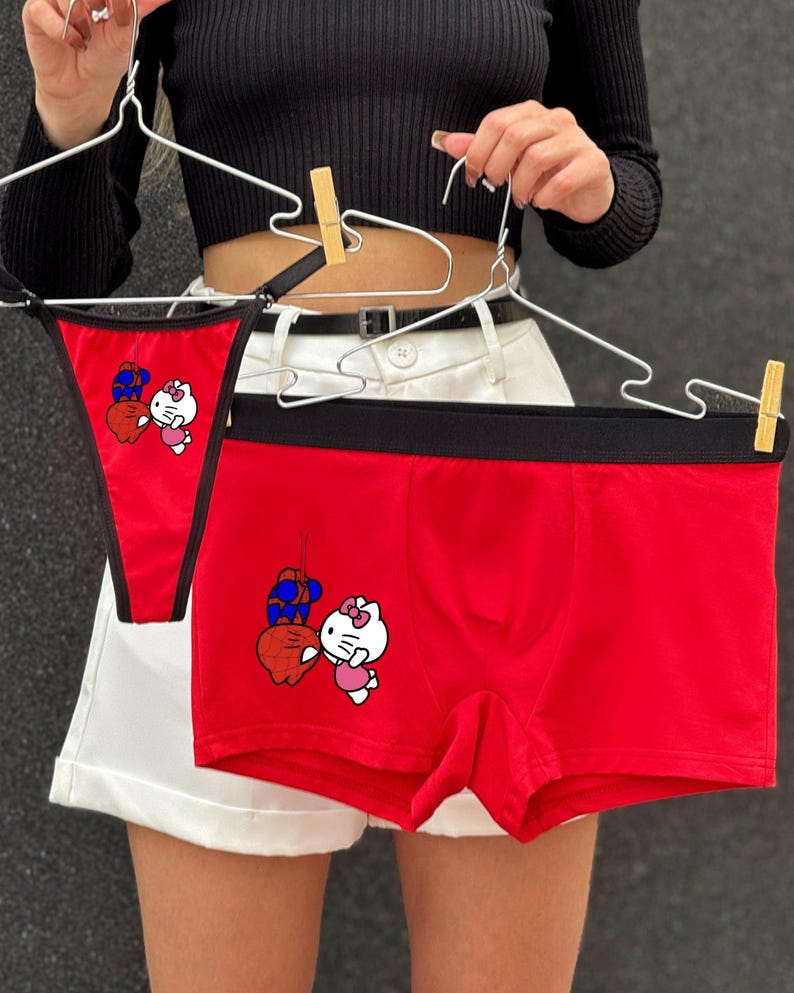 Pode incluir: Conjunto de roupa interior vermelha com fio dental e cuecas boxer. Ambas as pe&ccedil;as t&ecirc;m uma cintura preta e um desenho animado de um gato branco com um la&ccedil;o rosa e um objeto azul e vermelho. A roupa interior est&aacute; pendurada em cabides de arame.