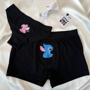 Puede incluir: Conjunto de ropa interior negra con personajes de dibujos animados. El conjunto incluye un bóxer con un personaje azul y una braga con un personaje rosa. En la imagen también hay una caja de regalo blanca con una cinta y una tarjeta.