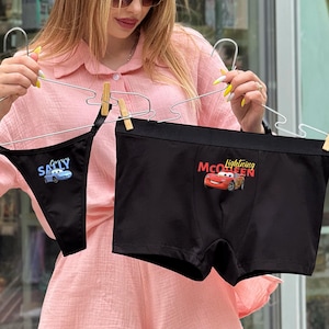 Puede incluir: Ropa interior negra con un gráfico de Lightning McQueen y un tanga negro con un gráfico de Sally. Ambos se muestran en perchas. La ropa interior es negra con un gráfico de Lightning McQueen y el tanga tiene un gráfico de Sally.