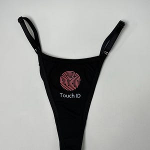 May include: A black thong featuring a pink circular fingerprint graphic centred on the front. Below the graphic, the text "Touch ID" is printed in white. The garment includes thin, adjustable black shoulder straps with metal sliders.