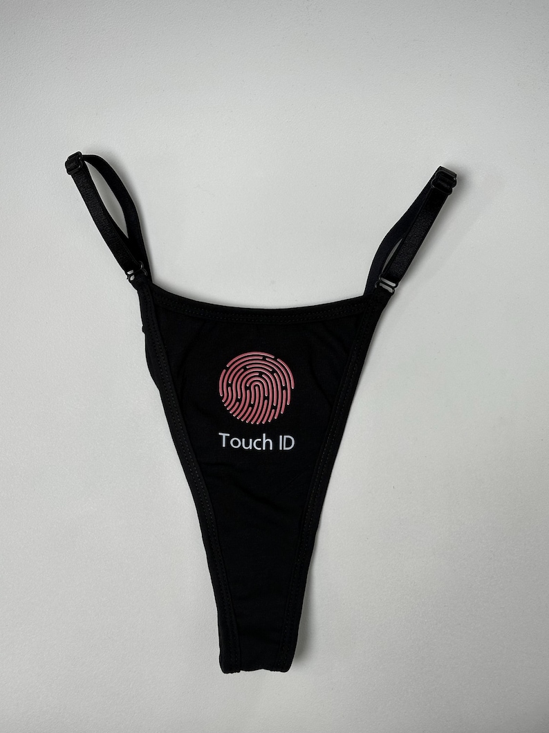 May include: A black thong featuring a pink circular fingerprint graphic centred on the front. Below the graphic, the text "Touch ID" is printed in white. The garment includes thin, adjustable black shoulder straps with metal sliders.