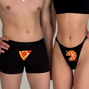 Op de afbeelding: Zwarte boxershorts en een zwarte string, beide met pizzapuntontwerpen. De boxershorts hebben een enkele punt, terwijl de string een hele pizza met een ontbrekende punt toont. De pizzapunten hebben tomaten-, kaas- en peper toppings.