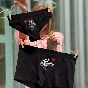 Könnte beinhalten: Schwarze Unterwäsche und Boxershorts mit einem Cartoon-Schrauben- und Muttern-Design. Die Unterwäsche hat einen kurzen Schnitt, und die Boxershorts haben eine längere, entspanntere Passform. Beide haben ein rotes Herz.