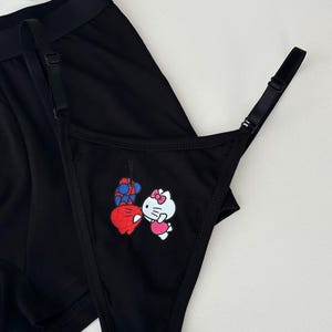 Pode incluir: Conjunto de roupa interior preta com fio dental e cuecas boxer. O fio dental tem uma estampa gr&aacute;fica de um personagem de desenho animado com uma fantasia vermelha e azul e um gato de desenho animado branco com la&ccedil;o e vestido rosa. As cuecas boxer s&atilde;o pretas lisas.