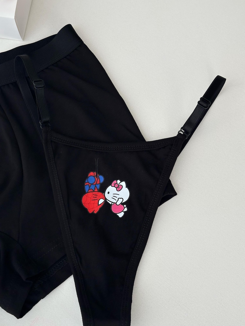 Pode incluir: Conjunto de roupa interior preta com fio dental e cuecas boxer. O fio dental tem uma estampa gr&aacute;fica de um personagem de desenho animado com uma fantasia vermelha e azul e um gato de desenho animado branco com la&ccedil;o e vestido rosa. As cuecas boxer s&atilde;o pretas lisas.