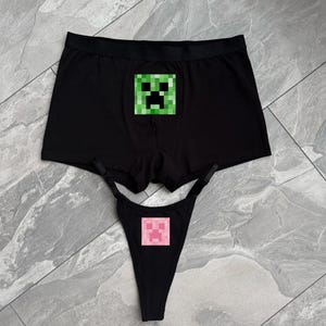 Könnte beinhalten: Eine schwarze Boxershorts mit einem grünen, verpixelten Creeper-Gesicht und ein dazu passender schwarzer Tanga mit verstellbaren Trägern, der auf der Vorderseite jeweils ein rosa, verpixelten Creeper-Gesicht als Grafik zeigt.