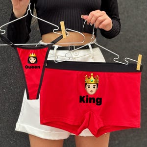 Könnte beinhalten: Rotes Unterwäsche-Set mit einem String mit dem Text "Queen" und einer Boxershorts mit dem Text "King", beide mit Kronen-Emojis. Das Set wird an Kleiderbügeln präsentiert.