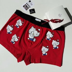 Könnte beinhalten: Rote Boxershorts mit schwarzem Bund, bedruckt mit Cartoon-Bildern von Hello Kitty, die den Mittelfinger zeigen. Eine weiße Geschenkbox mit rotem Band und dem Text "TRENDY PRINT" ist ebenfalls zu sehen.