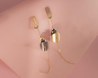 May include: Two contemporary earrings in gold and silver. Each earring has a unique design, combining geometric shapes like ovals, rectangles, and rounded elements. They hang from slender, curved gold wires, set against a pink backdrop.