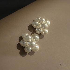May include: Two clusters of white faux pearl earrings. Each earring features multiple pearls of varying sizes, arranged to resemble a small bunch. The earrings are set against a neutral, light brown background.