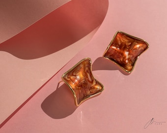 May include: A pair of gold-toned earrings with a geometric, square-like shape. The earrings are made of a brown, marbled material with gold accents.