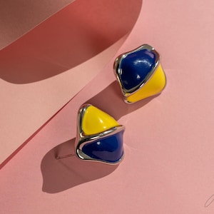May include: A pair of blue and yellow enamel earrings with a silver metal setting. The earrings are shaped like a teardrop with a curved edge.