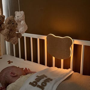May include: A crib mobile featuring four plush teddy bears in white and brown, suspended from a circular frame. A bear-shaped wooden wall light with a warm glow is mounted on the white crib railing. The crib sheet has a small brown bear pattern.