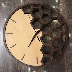 May include: A round wooden wall clock with a unique design. The clock face is divided into two sections: a light wood half and a dark wood honeycomb pattern. Black clock hands and minimalist hour markers complete the design.