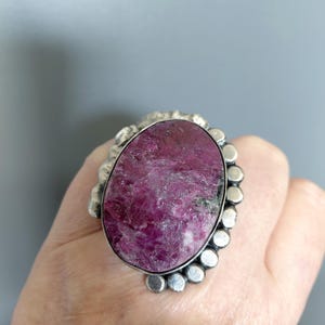 May include: A large, oval-shaped ring featuring a vibrant, textured, magenta-colored gemstone. The gemstone is set in a silver-toned metal with a decorative, beaded border. The ring is a statement piece, showcasing a bold and eye-catching design.