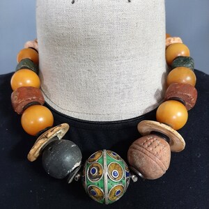 May include: A colorful beaded necklace with a variety of materials, including amber, wood, and metal. The necklace features a large, round, green and gold bead with intricate patterns.