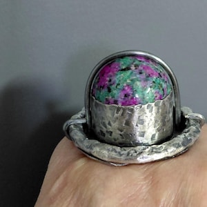 Puede incluir: Anillo artesanal con una piedra preciosa grande y redonda con una mezcla de colores rosa, verde y negro. La piedra está engastada en un engaste de metal plateado martillado con un diseño único y artístico. El anillo es una joya llamativa.