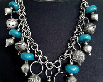 May include: A silver chain necklace with turquoise blue beads and silver charms. The necklace has a variety of charms, including bells, rings, and round beads.