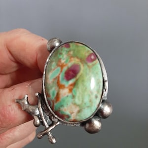 Może przedstawiać: Srebrny pierścionek z zielonym i czerwonym kamieniem osadzonym w srebrnym owalu. Pierścionek ma srebrny wzór jelenia po bokach.