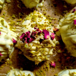 May include: A group of egg-shaped confections coated in white chocolate, drizzled with additional white chocolate, and topped with crushed green pistachios and dried pink rose petals, arranged on a brown surface.