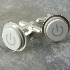 Pode incluir: Um par de botões de punho de prata com um design de botão de energia branco. Os botões de punho são redondos e têm uma borda prateada.
