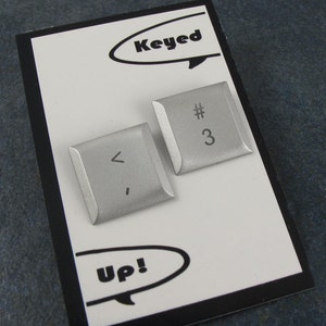 Peut inclure: Deux touches de clavier en métal argenté. Les touches sont de forme carrée et ont des coins arrondis. Les touches sont gravées avec les symboles "<" et "#3".
