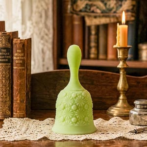 May include: A pale green glass bell with a textured handle and a patterned body. The bell is displayed on a wooden surface with antique books and a lit candle in the background. The bell is approximately 6 inches tall.