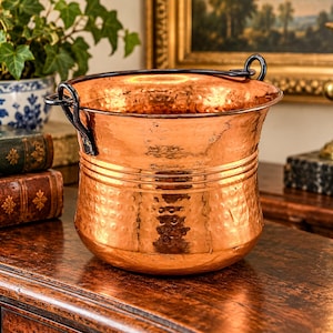 May include: A round copper bucket with a hammered texture on the lower half and a smooth upper rim. It features a dark metal swing handle attached to two side loops. The vessel has a wide opening and a slightly tapered base, showcasing a metallic finish.