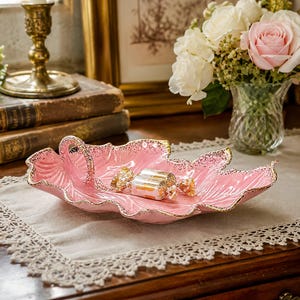 May include: A pink ceramic serving dish shaped like a large leaf with a textured surface and a gold-painted scalloped rim. The dish features a small integrated handle on one side and contains a single wrapped candy in the center of the bowl.