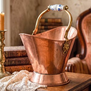 May include: A copper bucket with a brass handle and decorative accents. The handle features a white and blue ceramic grip. The bucket sits on a wooden table, with a candle and books in the background. The bucket is approximately 10 inches tall.