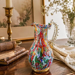 May include: A glass pitcher featuring a vibrant millefiori pattern with multicolored floral shapes in red, blue, green, and yellow. The pitcher has a clear, textured, spine-like handle and a gold-toned base, showcasing a rounded body and a flared spout.